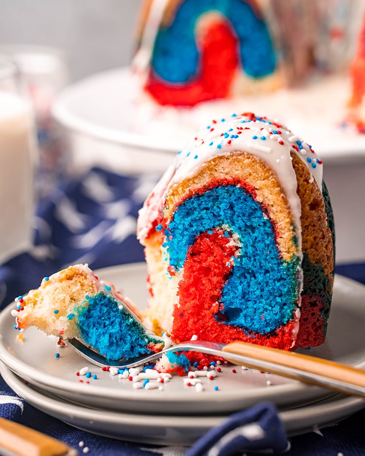 A slice of red white and blue bundt cake on a plate topped with a white glaze and patriotic sprinkles, with a fork taking a piece off the cake.