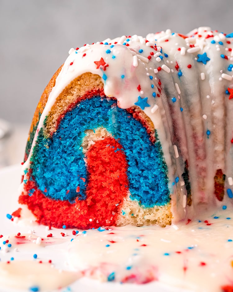 A cut of a patriotic bundt cake that is showing the red, white, and blue colors topped with a white glaze, and red, white, and blue sprinkles.