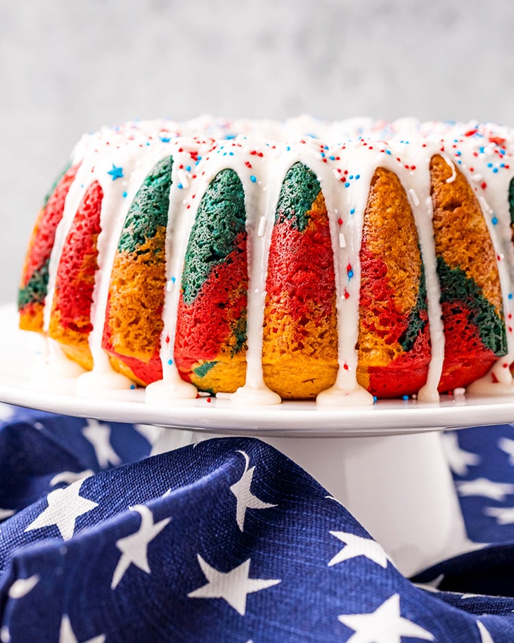 A bundt cake on a white stand, the cake has red, blue, and yellow colors. There is a white glaze over the top with red and blue sprinkles.