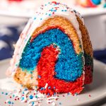 A slice of red white and blue bundt cake on a plate topped with a white glaze and patriotic sprinkles.