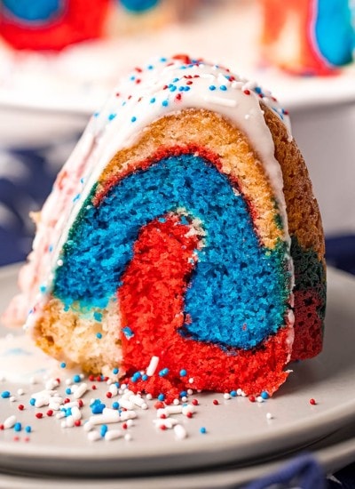 A slice of red white and blue bundt cake on a plate topped with a white glaze and patriotic sprinkles.