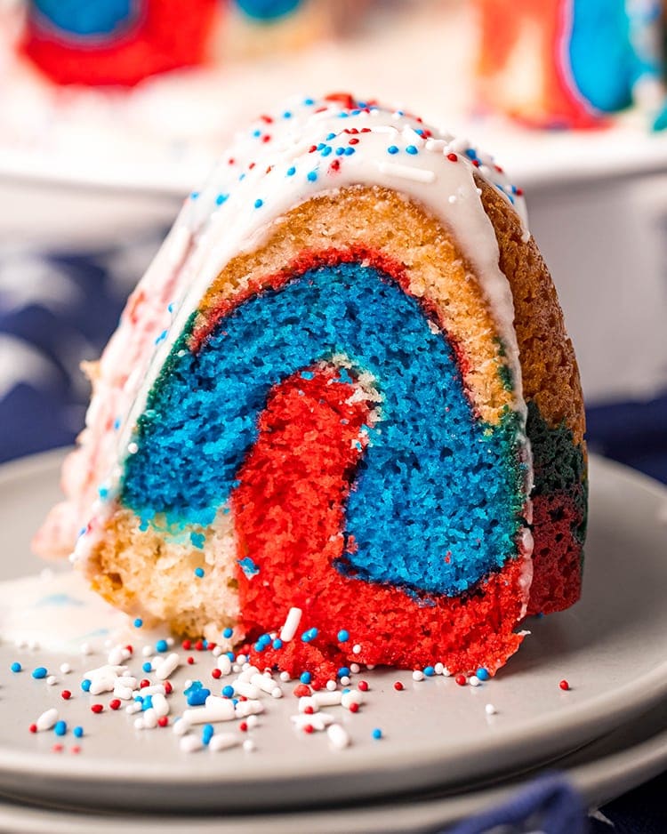 A slice of red white and blue bundt cake on a plate topped with a white glaze and patriotic sprinkles.