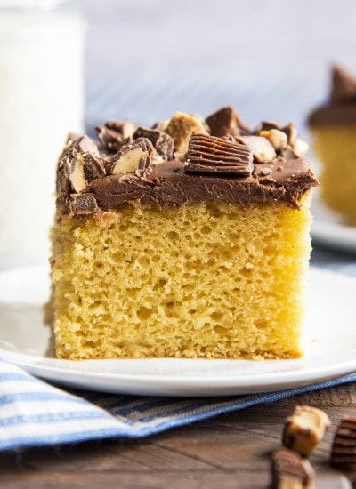 A square piece of peanut butter cake on a white plate topped with chocolate frosting and chopped peanut butter cups.