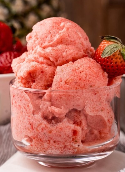 A clear glass bowl piled high with a pink strawberry pineapple sorbet, topped with a fresh strawberry.