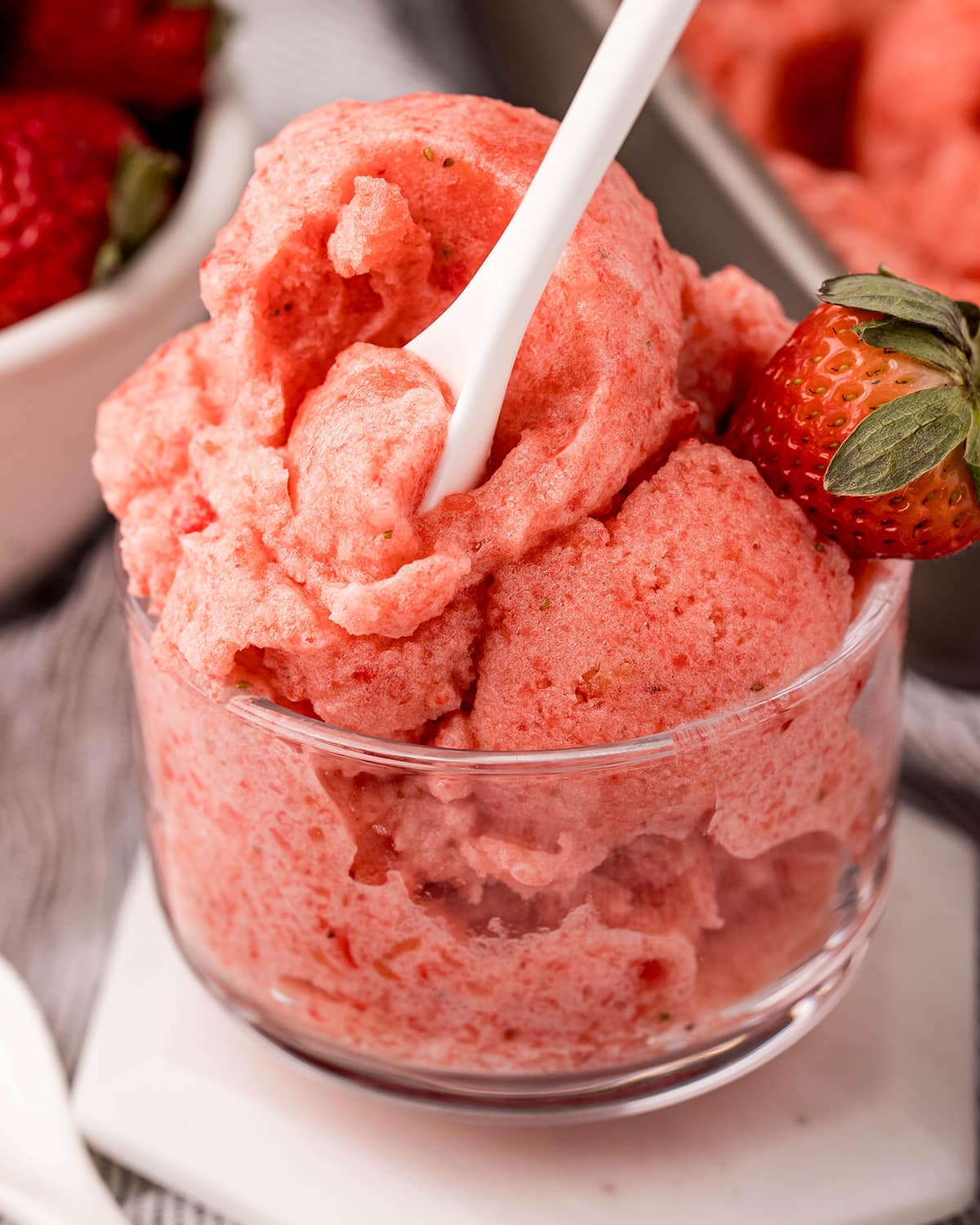 A glass bowl of strawberry sorbet with a small white spoon holding cutting into the scoop.