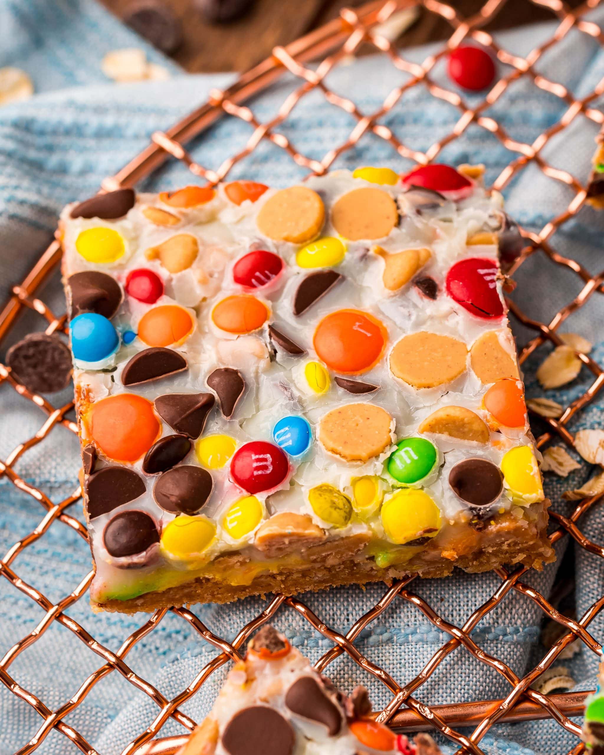 A close up of a monster cookie magic bar on a wire rack, with a variety of m&m colors, chocolate chips, and peanut butter chips in a white sweetened condensed milk layer.