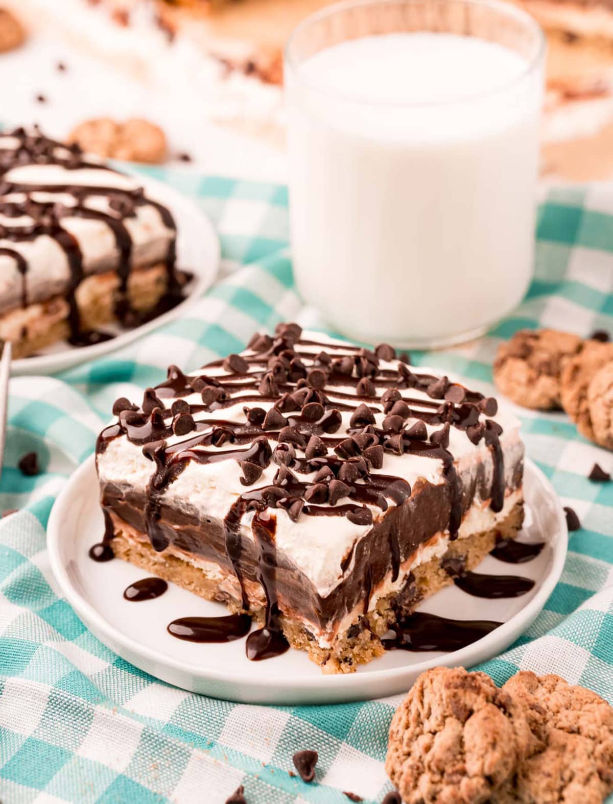A slice of a layered chocolate chip cookie delight on a small plate. It has a layer of cookie, cream cheese mixture, chocolate pudding, cool whip, and is topped with mini chocolate chips and chocolate syrup.