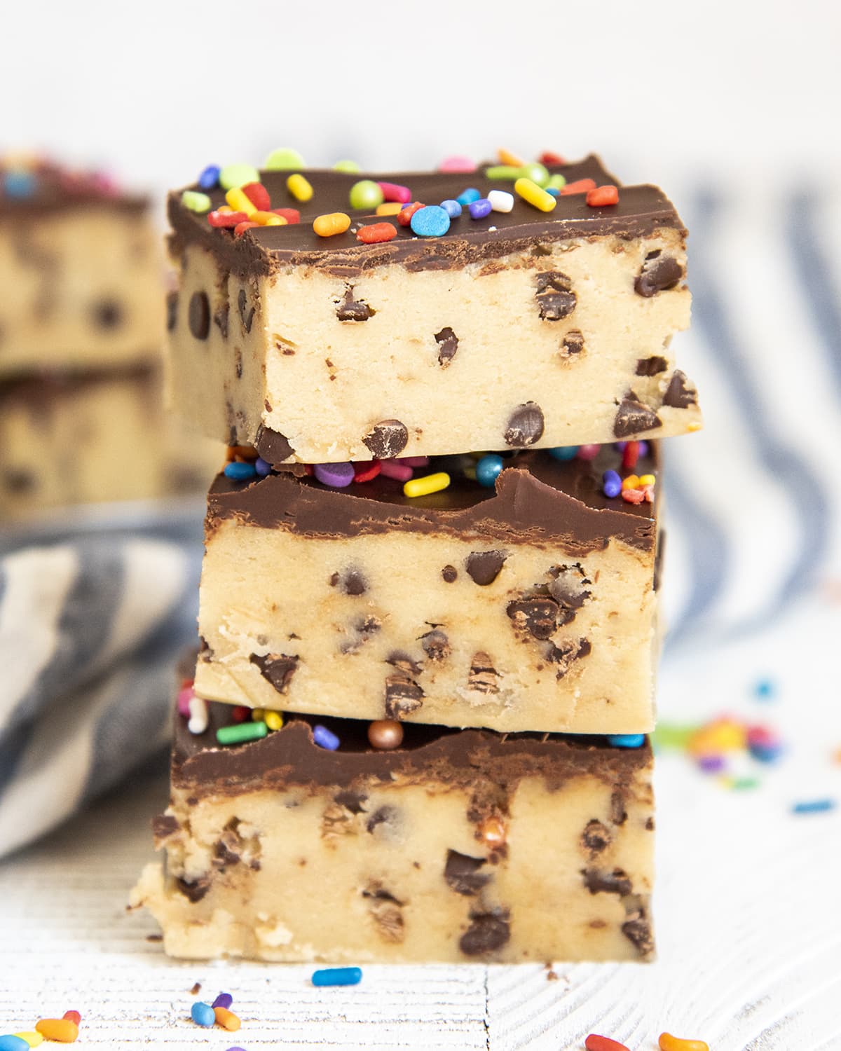 A stack of bars that are made of cookie dough filled with mini chocolate chips, and topped with a layer of chocolate, and sprinkles.