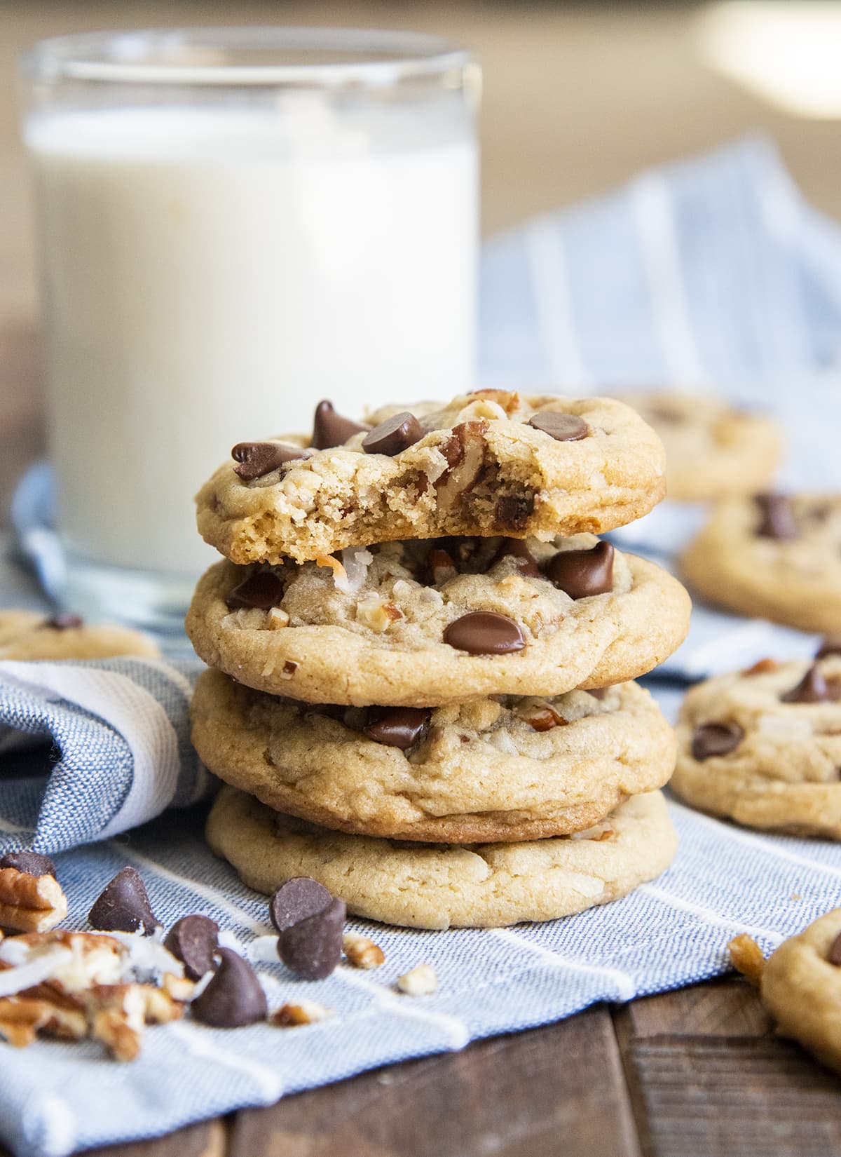 A stack of 4 drop style cookies with chocolate chips, pecans, and shredded coconut. The top cookie has a bite taken out of it.