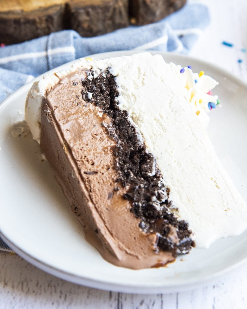 A slice of ice cream cake laying on a plate, the bottom half is chocolate, with a layer of oreo crumbs, then vanilla ice cream, and topped with whipped cream and a whipped rosette.