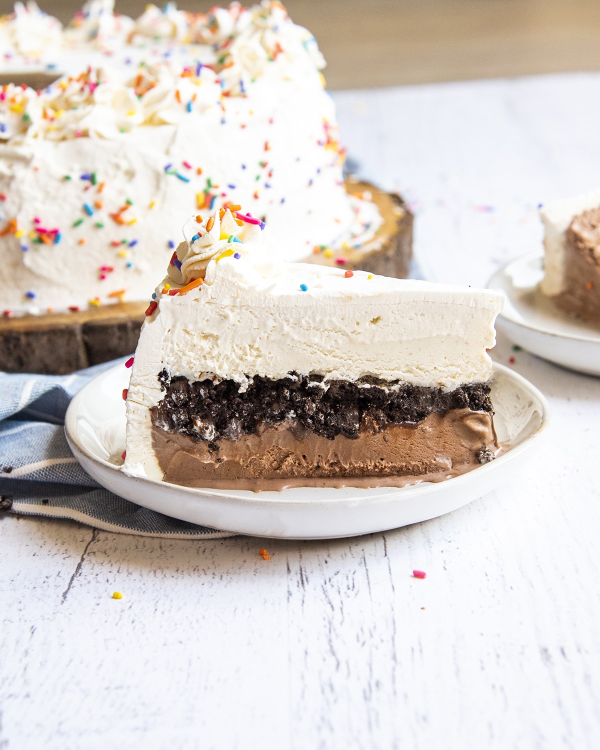 A piece of ice cream cake standing on a plate. The cake has chocolate ice cream then oreo crumbs, then vanilla icecream, and whipped cream on the outside and top like frosting.