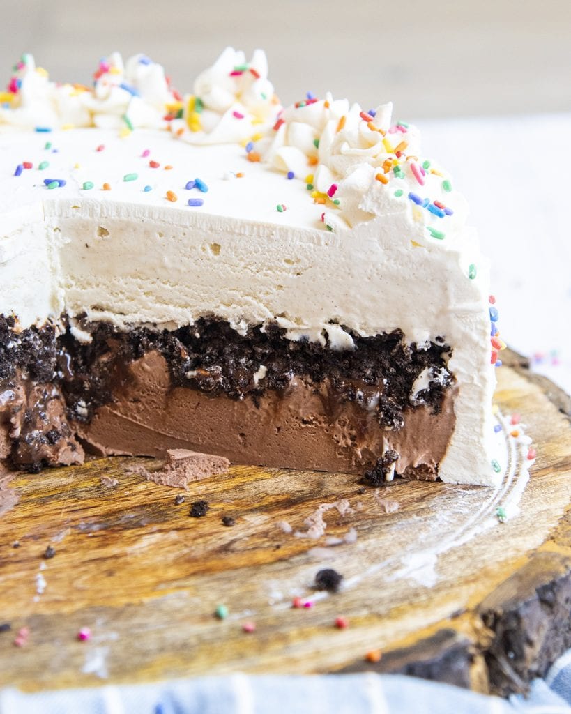 An ice cream cake on a wooden board, cut open to reveal the inside of the cake with chocolate ice cream, oreo crumbs, and vanilla ice cream on top.