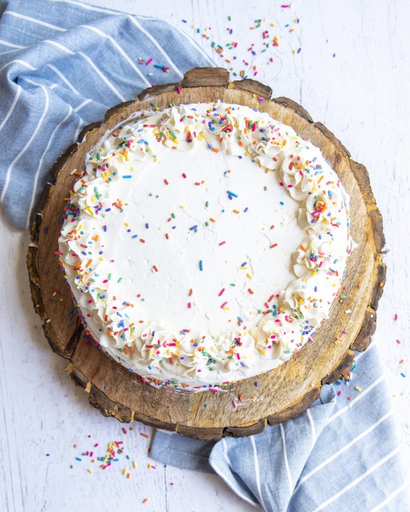 A white cake on a wooden round board, topped with swirls of frosted rosettes and sprinkles.