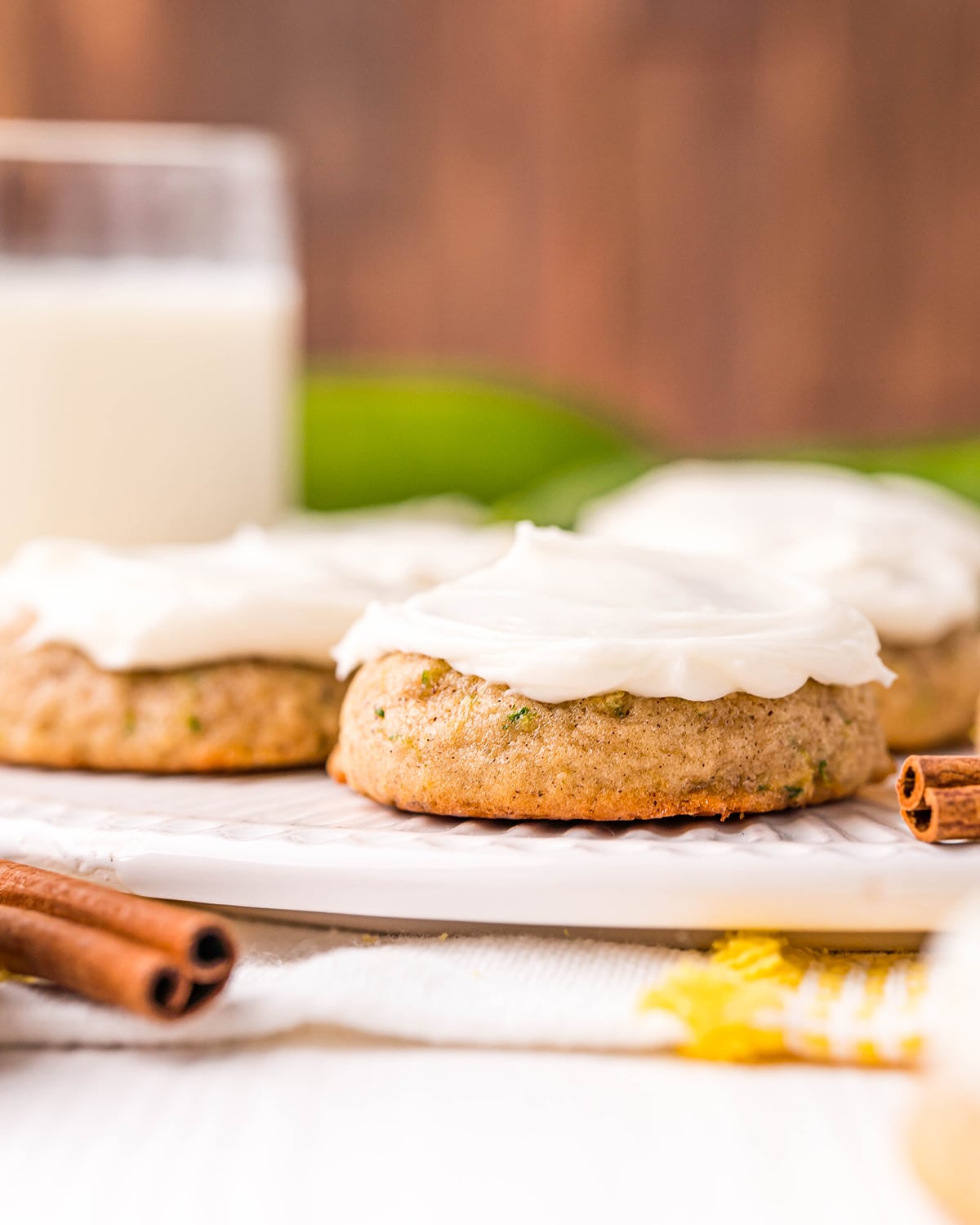 Zucchini cookies on a white stand with zucchinies in the back, and a cup of milk.