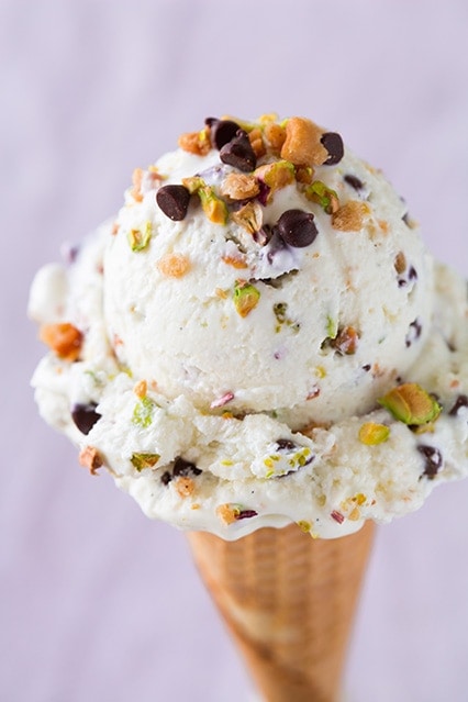 An ice cream cone topped with vanilla ice cream with pistachio pieces, and mini chocolate chips throughout and on top of the scoop.