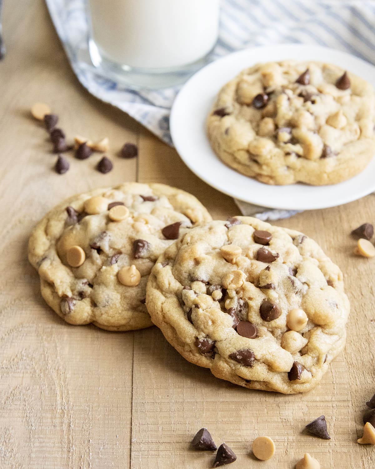 Two giant cookies full of chocolate chips and peanut butter chips on a wooden surface, with another cookie on a plate behind them.