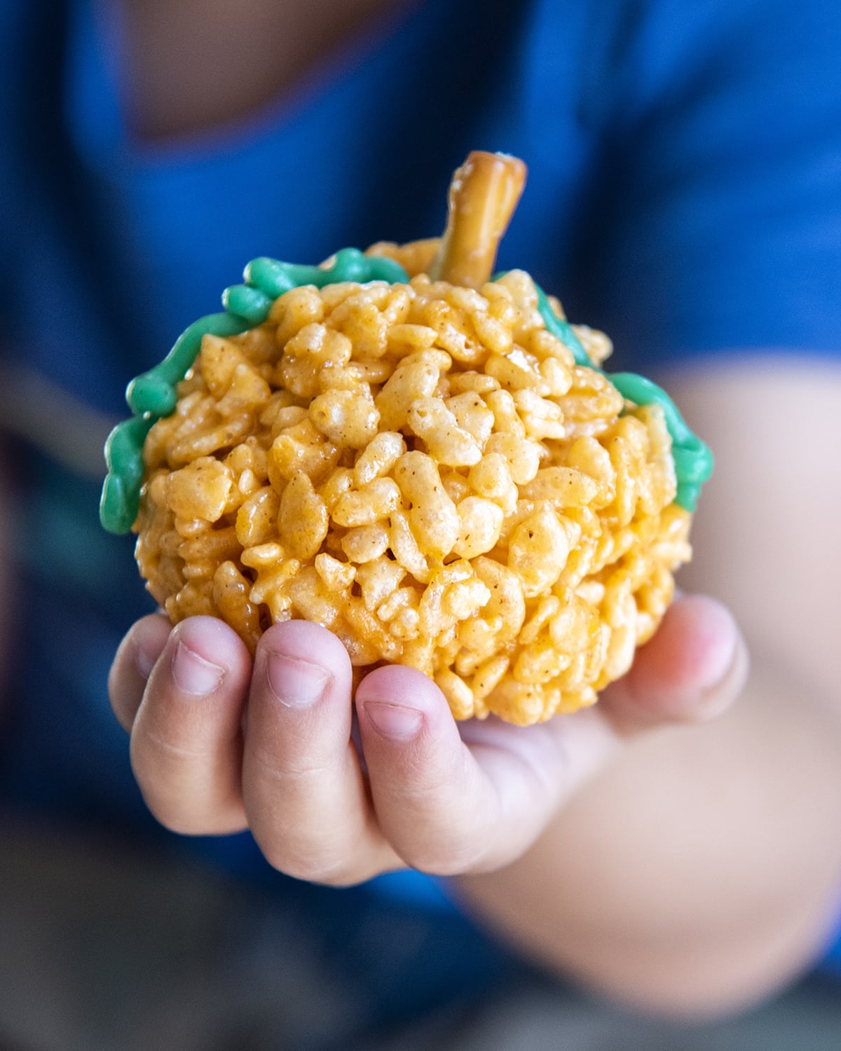 A child's hand holding a rice krispie treat that is shaped and colored to look like a pumpkin.