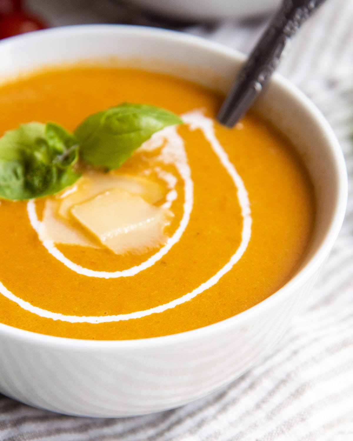 A close up on a bowl of tomato soup topped with a swirl of heavy cream, and parmesan cheese, and basil leaves.