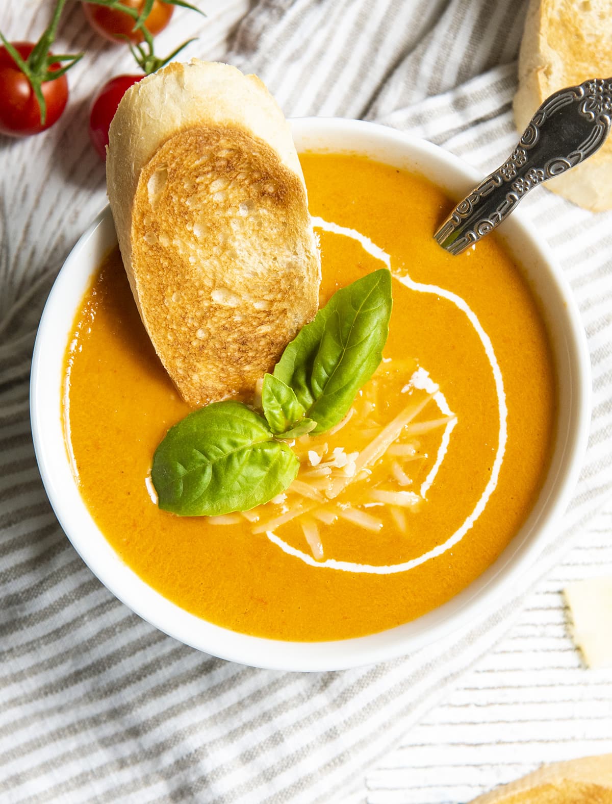 An overhead shot of a bowl of roasted tomato soup topped with shredded parmesan, fresh basil, and a piece of toasted bread. 