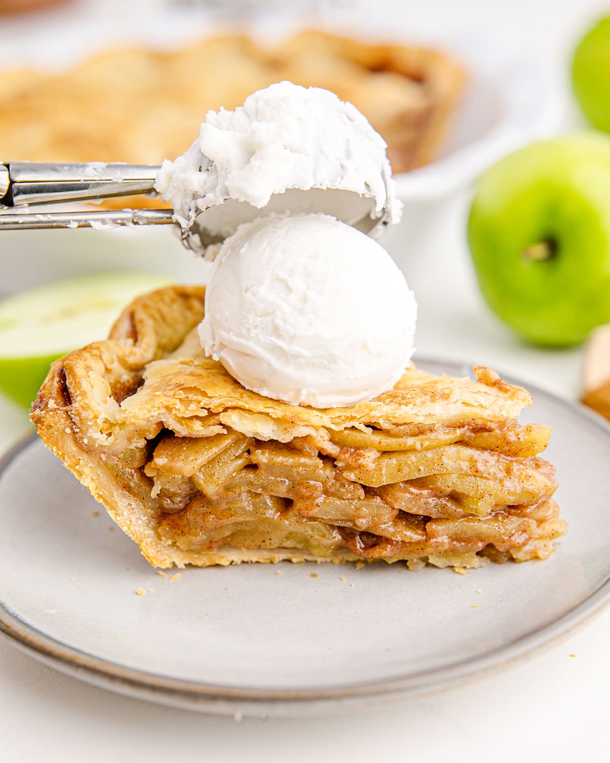 A piece of apple pie on a plate topped with a scoop of ice cream coming out of the ice cream scooper.