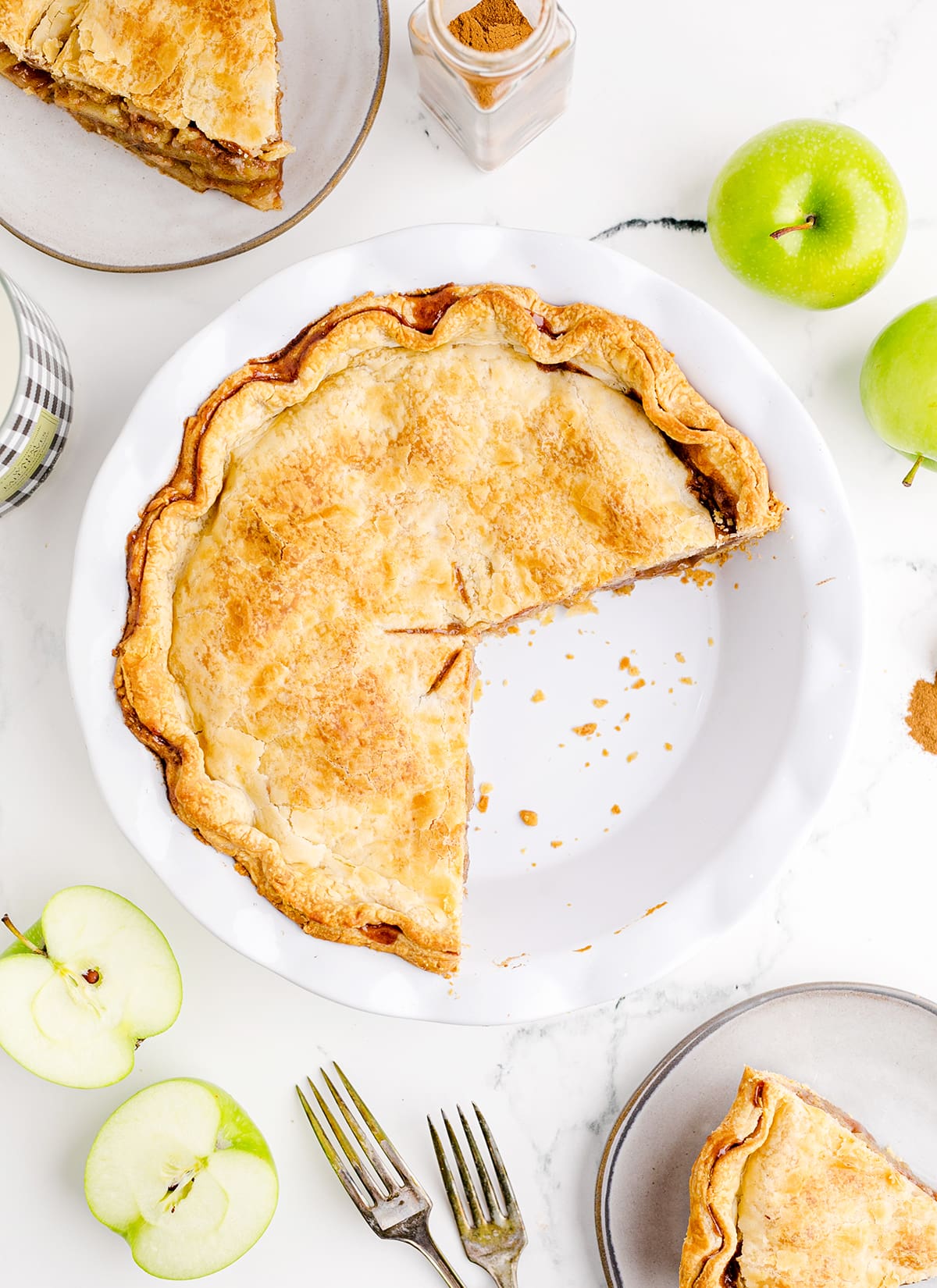 An overhead photo of 2/3 of an apple pie in a white pie plate with apple pie slices and apples surrounding it.