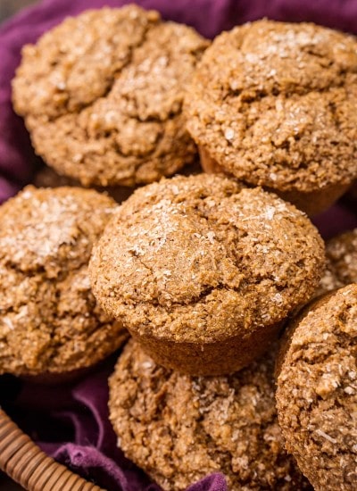 A pile of bran muffins in a basket lined with a purple cloth.