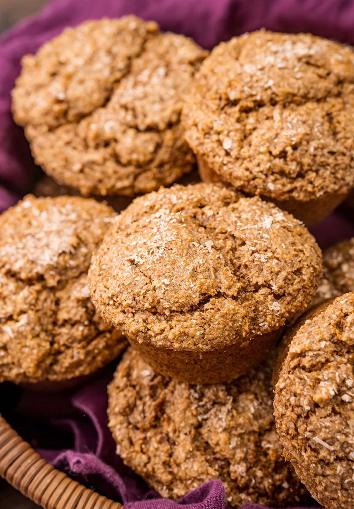 A pile of bran muffins in a basket lined with a purple cloth.