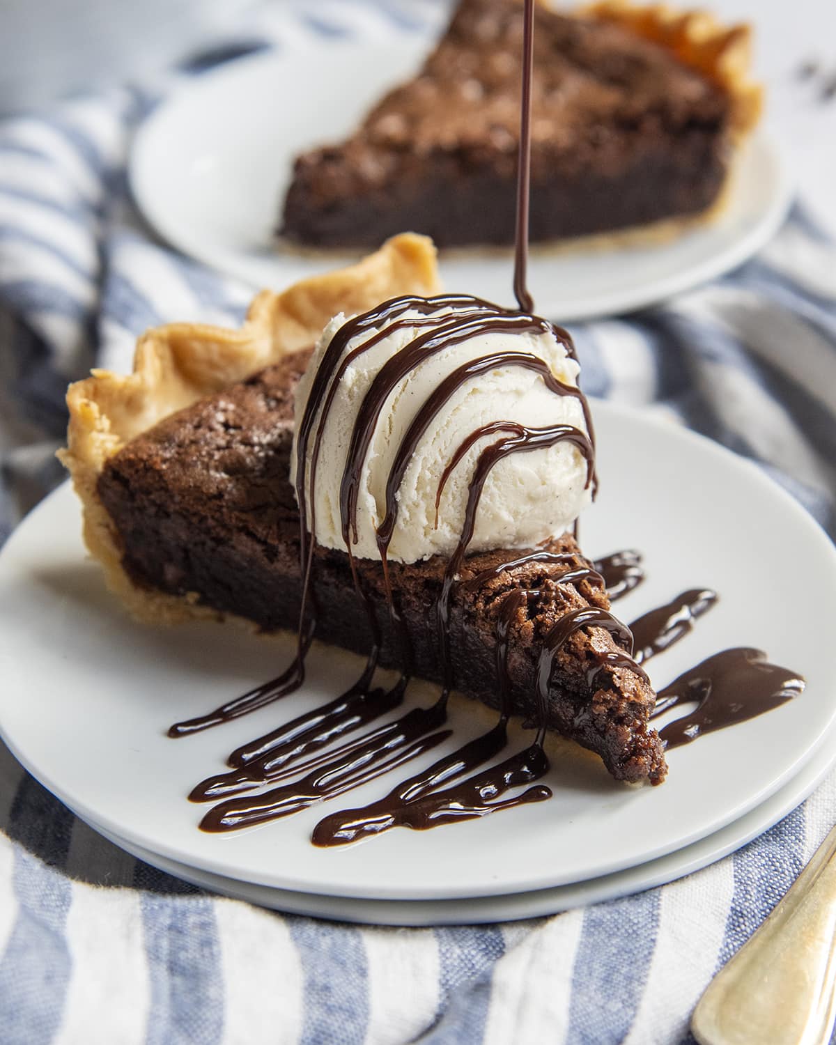 A slice of brownie pie on a plate, topped with ice cream, and chocolate sauce being drizzled over them both.