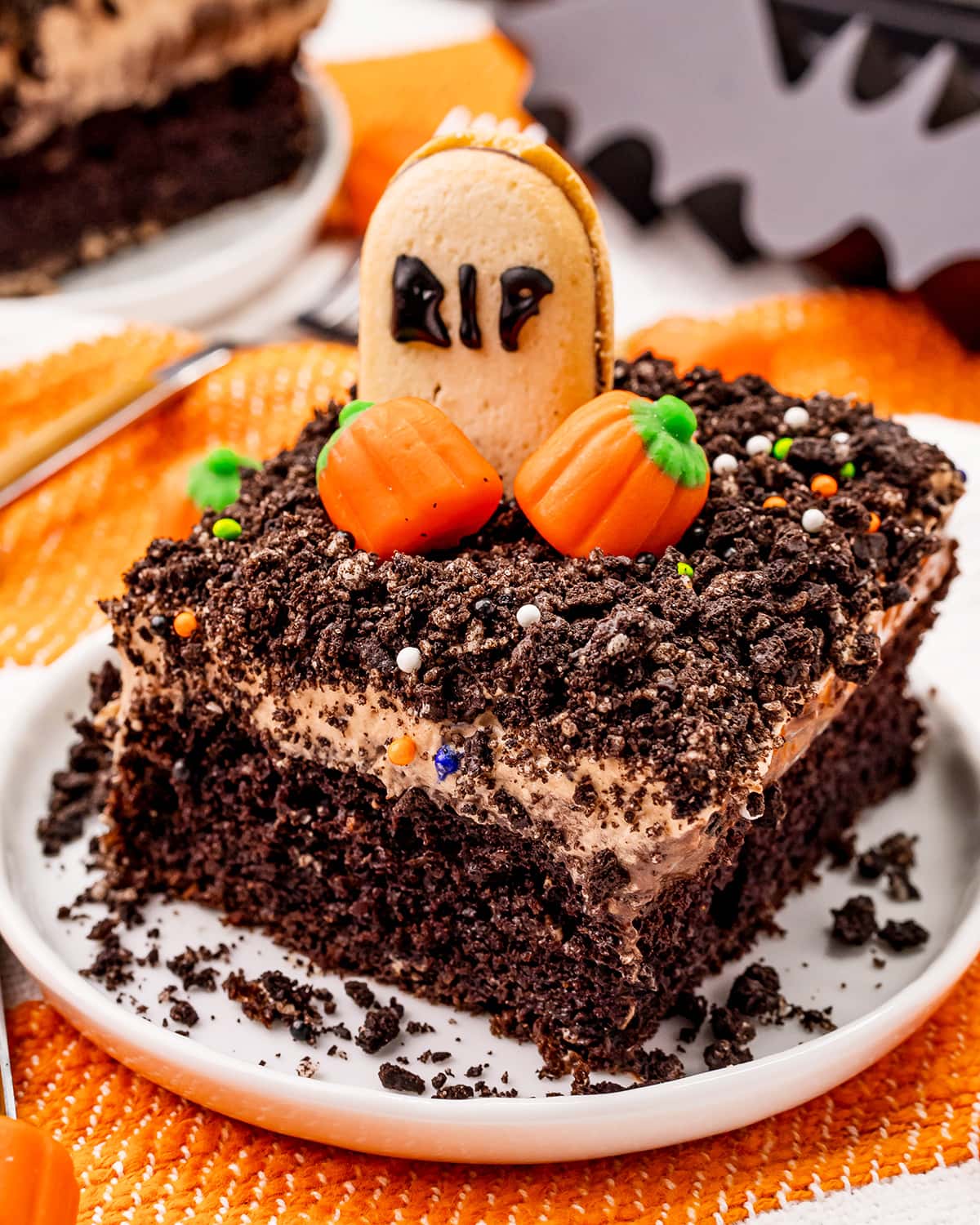A piece of a chocolate cake topped with chocolate frosting, Oreo crumbs, candy pumpkins, and a Milano cookie with the letters RIP written on it.