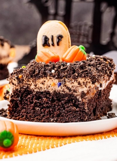 A slice of a Halloween Dirt Cake on a plate topped with a "gravestone" cookie and candy pumpkins, and Oreo crumbs.