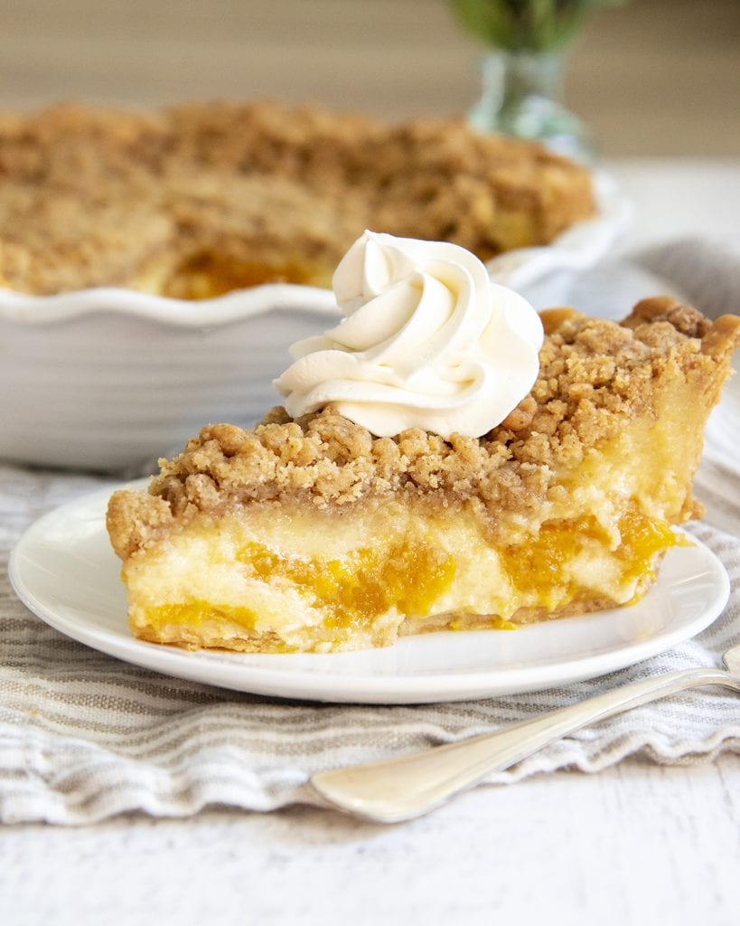 A side shot of a slice of sour cream peach pie, showing the custard, and slices of peaches, and crumble topping on top. With a swirl of whipped cream.