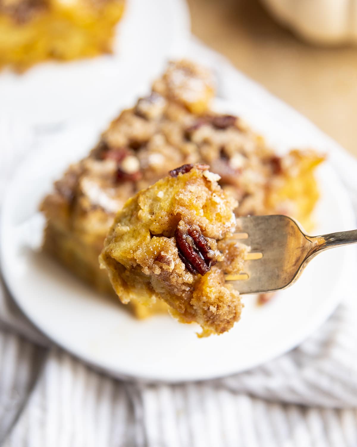 A bite of french toast casserole on a fork above a serving of it on a dish. 