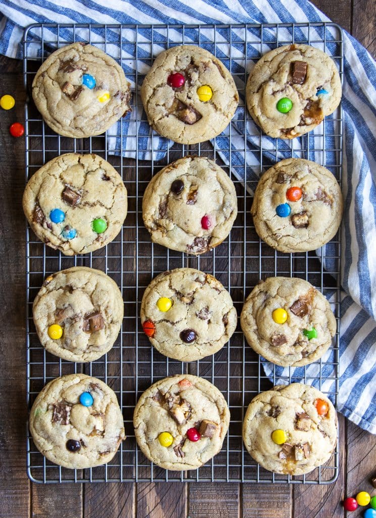 A cooling rack lined with 12 large cookies topped with a couple m&ms and chocolate candy bar pieces.