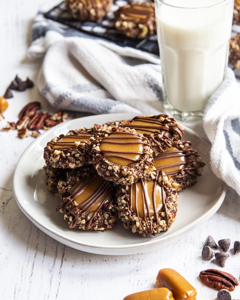 A plate of chocolate cookies coated in pecans, filled with caramel, and drizzled with chocolate.