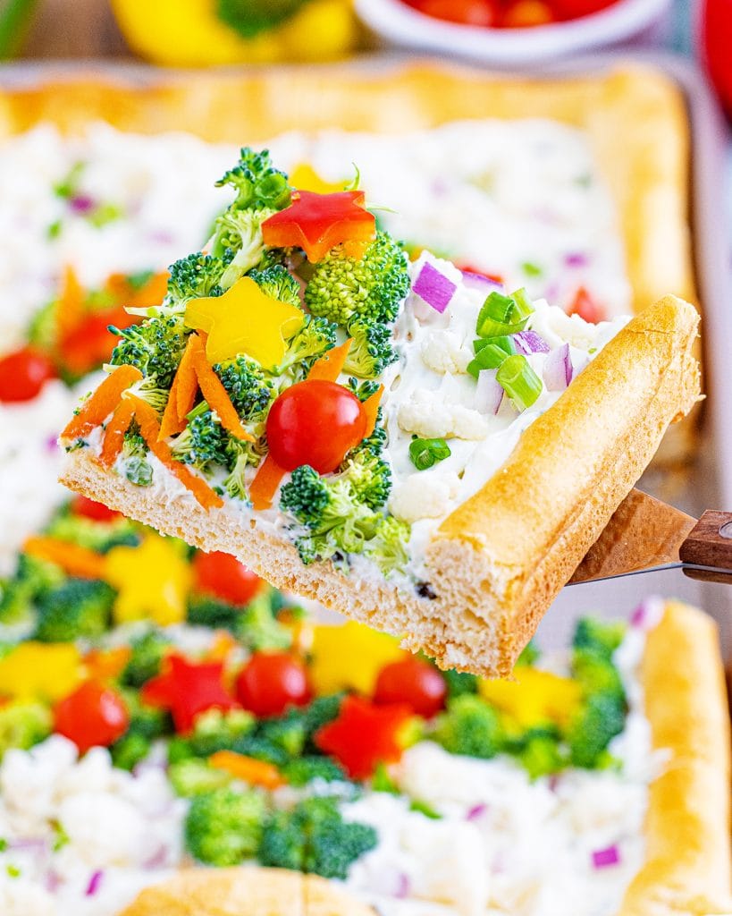 A piece of cold veggie pizza on a spatula topped with broccoli, shredded carrots, and half tomatoes.