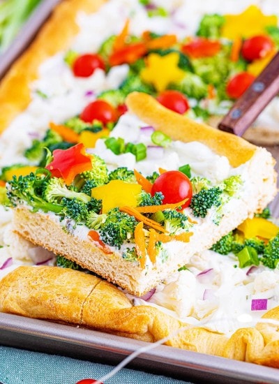 A piece of crescent roll veggie pizza on a spatula above a pan full of more pieces of the pizza.