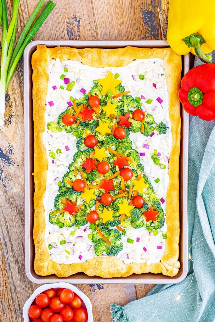 An overhead photo of a crescent roll veggie pizza in a pan, decorated with a broccoli christmas tree topped with tomatoes and pepper "ornaments"