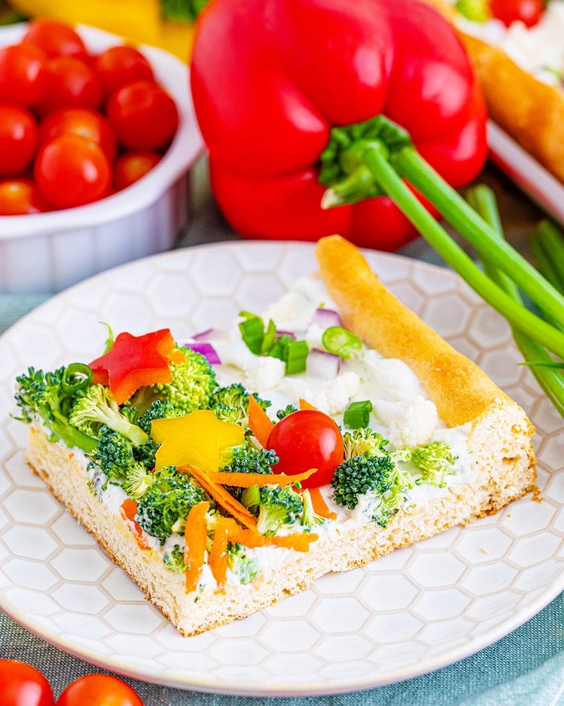 A piece of crescent roll veggie pizza on a white plate with tomatoes, and a bell pepper behing it.