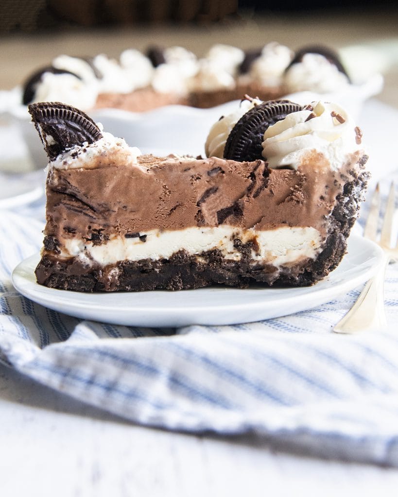A side shot of a piece of ice cream pie on a plate, with an Oreo crust, vanilla ice cream layer, chocolate ice cream layer, and whipped cream on top.
