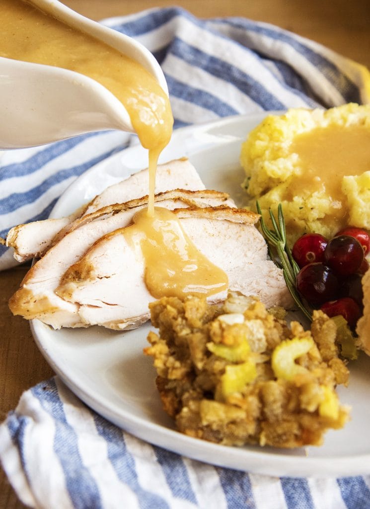 A plate of Thanksgiving food with mashed potatoes and gravy, stuffing, cranberries, and turkey breast slices with gravy being poured over the top.