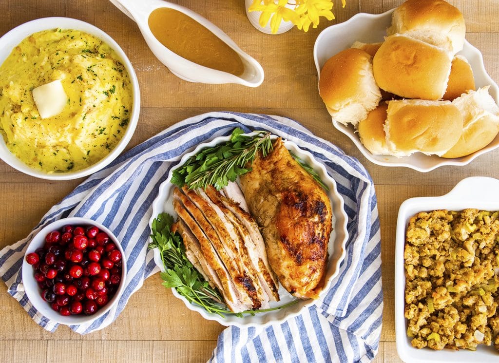 An overhead photo of a spread of Thanksgiving dishes. Turkey breast on a plate in the middle, surrounded by a bowl of rolls, baking pan of stuffing, mashed potatoes, and a pitcher of gravy.