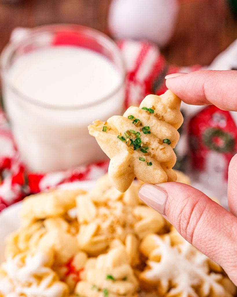 A hnad holding a small tree shaped buttery spritz cookie with green sprinkles. 