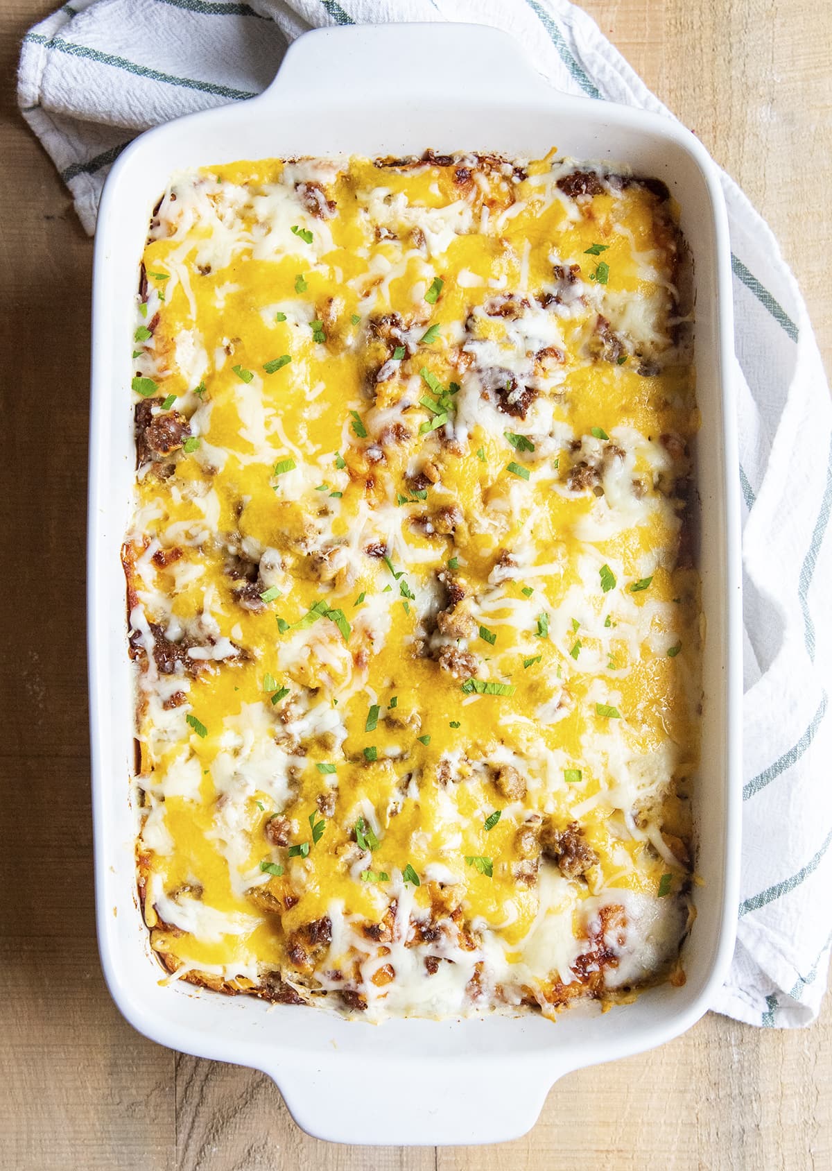 An overhead shot of a white 9x13 baking dish topped with cheese, pieces of sausage, and sprinkled with parsley.