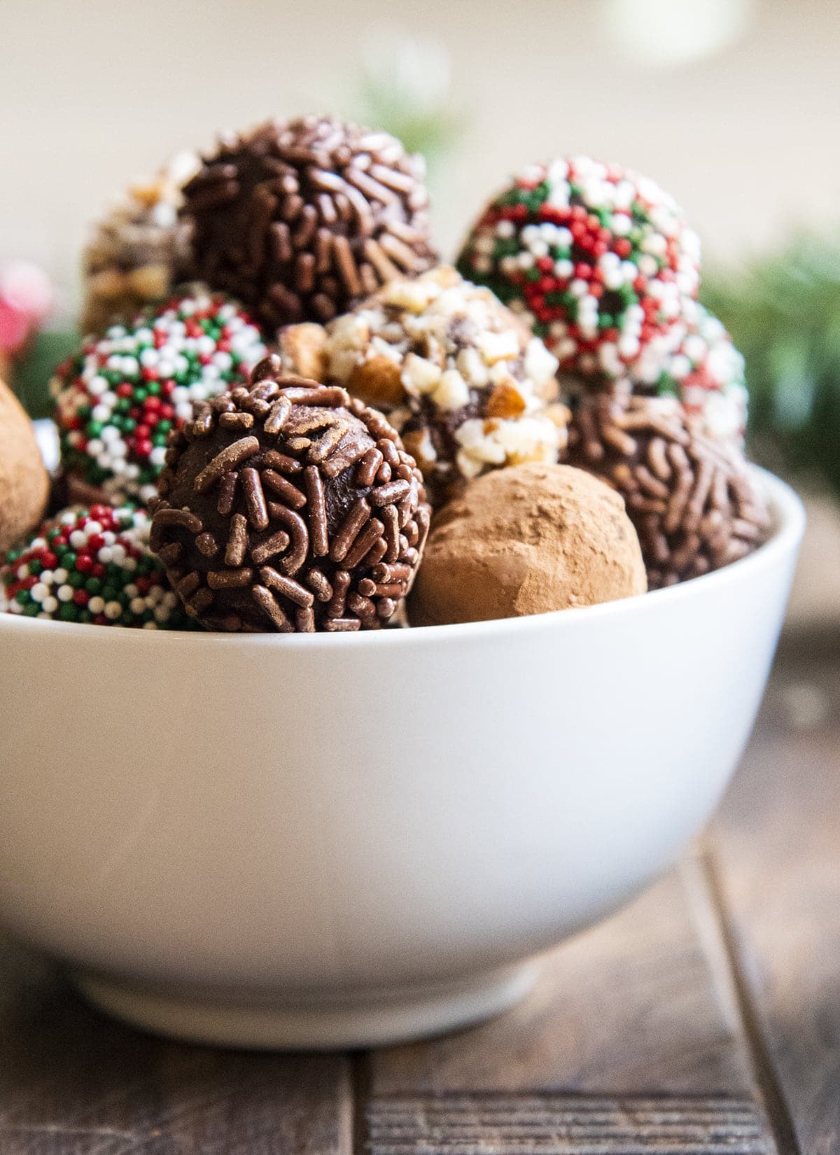 A white bowl full of chocolate truffles, some are coated in chocolate sprinkles, some in cocoa, some in chopped nuts.