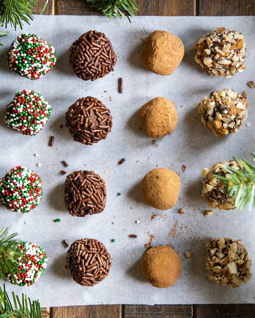 White parchment paper with chocolate truffles lined in rows of 4. Some are coated in Christmas sprinkles, some in chocolate sprinkles, some in cocoa powder, some in chopped almonds.