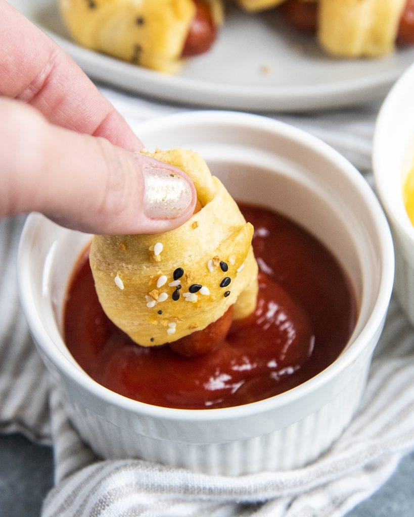 A hand holding a mini pigs in a blanket dipping it into ketchup.