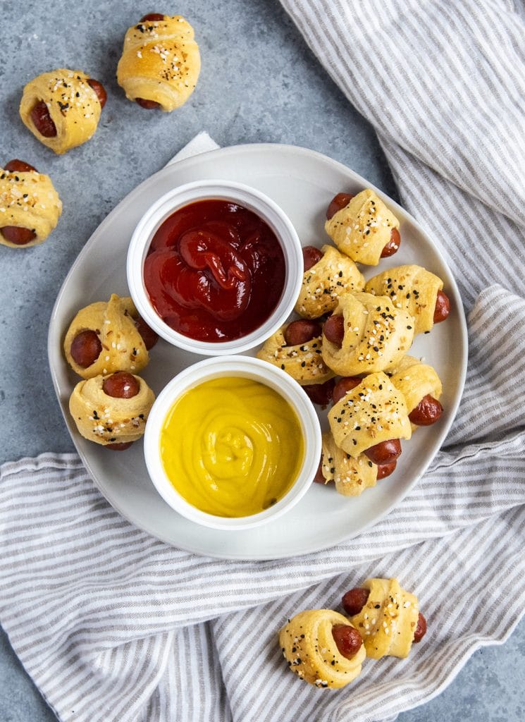 An overhead shot of a plate of pigs in a blanket with a bowl of ketchup, and a bowl of mustard.