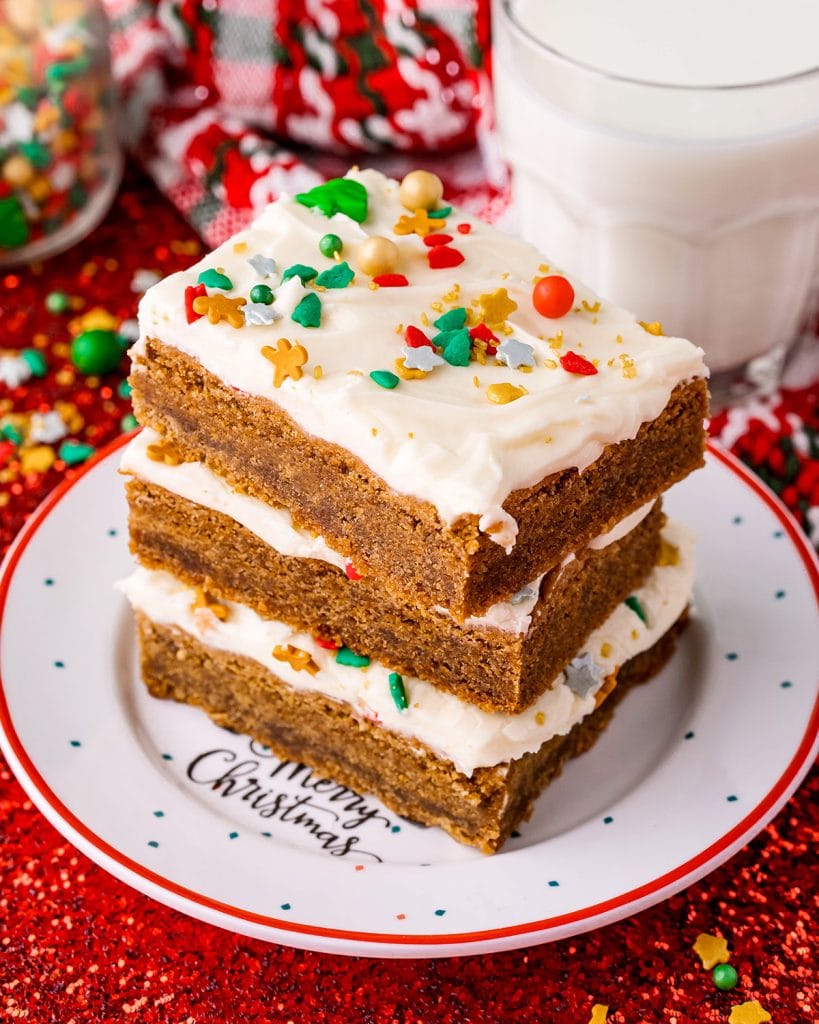 A stack of three ginger cookie bars topped with white frosting and holiday sprinkles on a holiday plate.