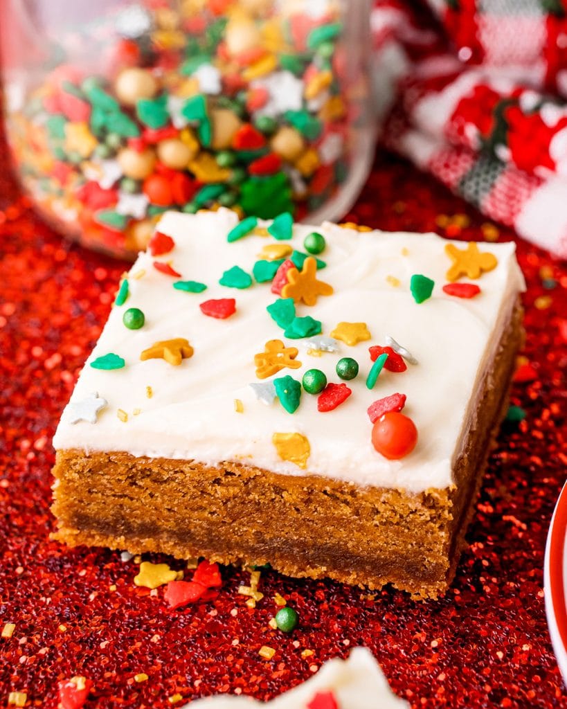 A gingerbread bar on a white sparkling background, the bar is topped with a white cream cheese frosting and red, gold, and green Christmas sprinkles.