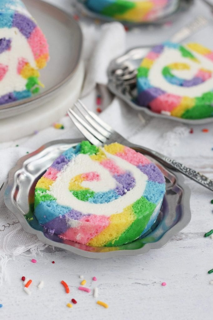 Slices of rainbow colored cake roll on silver plates.